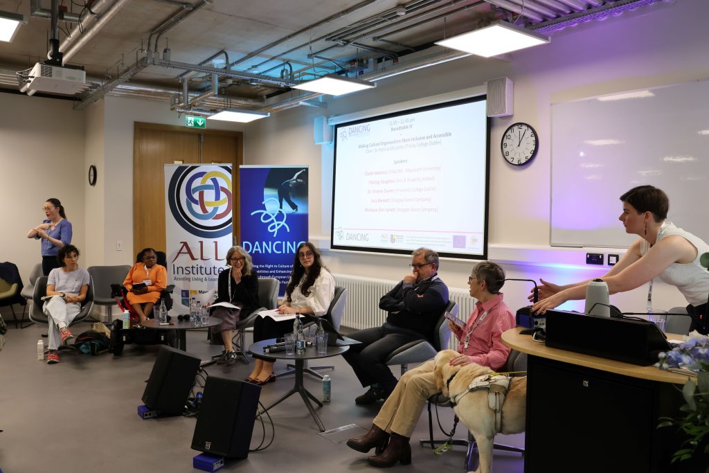 Third roundtable of the conference, featuring the chair and their guide dog, a member of the DANCING team helping to set up, the five speakers, and Irish Sign Language Interpretation.